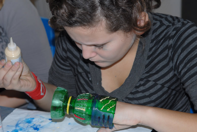 Na fotografiji je djevojka s Radionice antičko staklo. U ruci ima zelenu staklenu posudu koju ukrašava bojom.