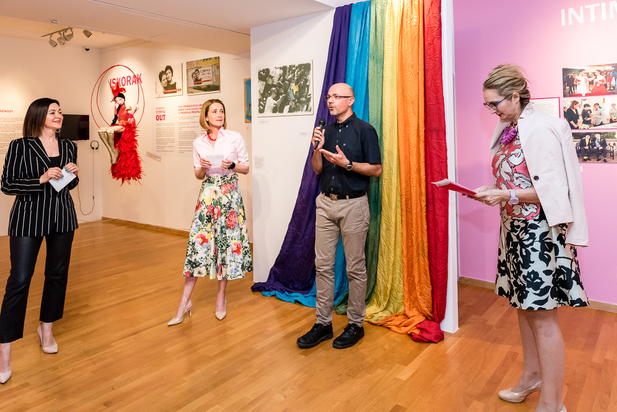 Na fotografiji je prikazan govor s otvorenja izložbe. Govore ravnateljica Tiflološkog muzeja Željka Bosnar Salihagić, Morana Vouk, Saša Tkalec i jedna ženska osoba. Saša Tkalec u ruci drži mikrofon, U pozadini se sa zida spušta platno u duginim bojama.