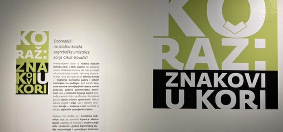 Na slici je postav izložbe. U desnom uglu je različitim veličinama slova napisano Koraž: znakovi u kori. Lijevo od naslova je legende naljepljena na zid. 